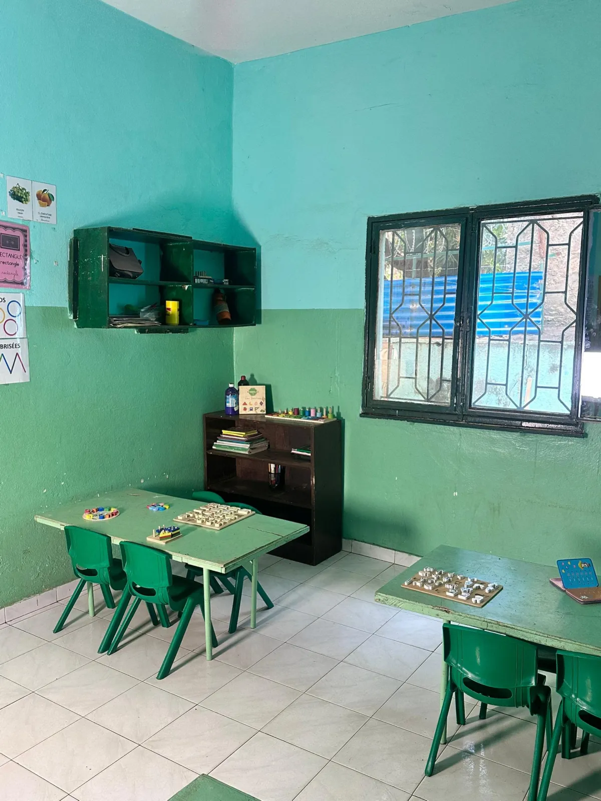 Salle de classe aux murs turquoise et vert : petites tables et chaises vertes, jeux éducatifs en bois et étagère de livres