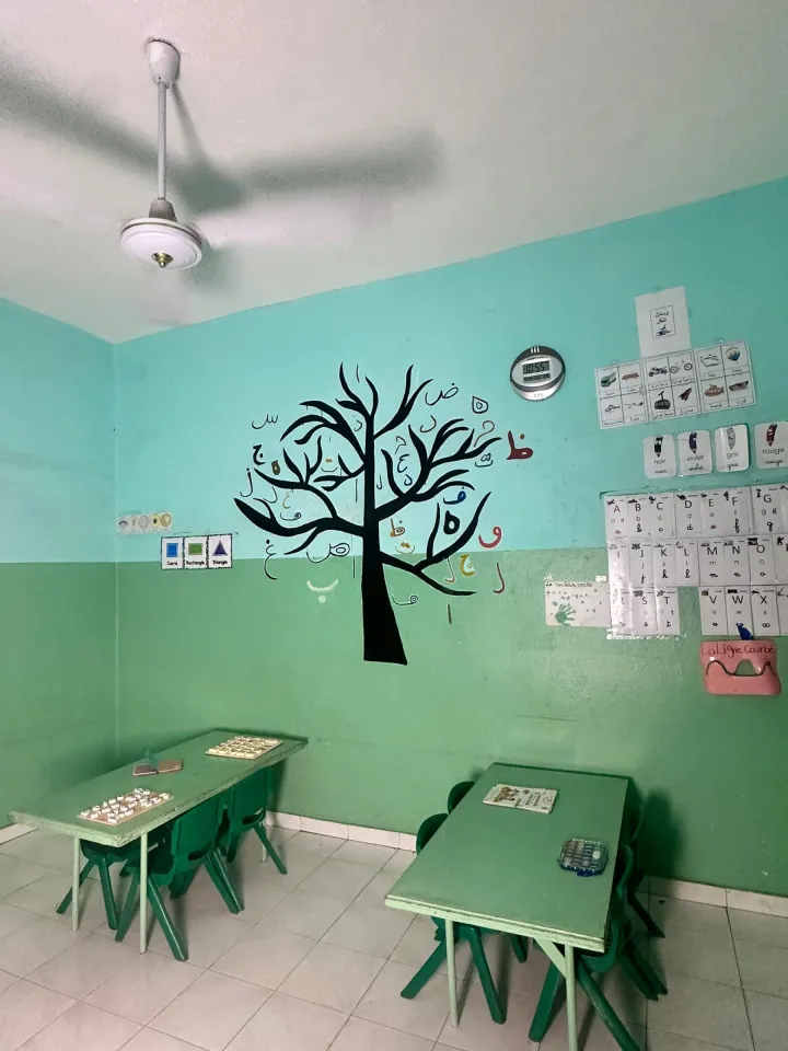 Vue d'ensemble d'une salle de classe : arbre de l'alphabet arabe, affiches en français et en arabe, tables et chaises vertes